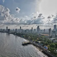 A wide drone shot of the Dadar coastline, capturing the viewing deck, the sea, and the expansive Mumbai skyline under a dramatic, sunlit sky.
