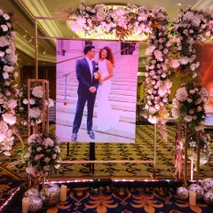 We create personalized walkways that tell your story. This entrance gallery features large, beautifully lit portraits of the couple, framed by elegant floral arrangements, allowing guests to walk through their journey of love.