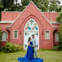A wide shot of a couple standing before a picturesque red chapel, with the mother-to-be's long blue gown spread out on the grass, creating a dramatic and romantic portrait.