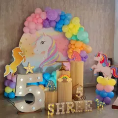 A magical unicorn-themed 5th birthday party. The centerpiece is a large unicorn backdrop surrounded by a pastel rainbow balloon garland, with unicorn cutouts and a light-up number '5'.
