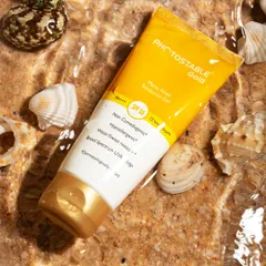 A creative shot of Photostable Gold sunscreen, styled in clear water with sand and seashells. This approach to skincare photography highlights the product's use case and creates a fresh, appealing visual for social media and ads.