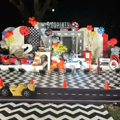 A complete race car-themed birthday stage. The setup features a road-patterned floor, a gas station prop, and large car cutouts, creating an immersive environment for a little car enthusiast's second birthday.