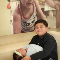 A proud big brother holds his newborn sibling for the first time. He smiles sweetly at the camera, showcasing the beginning of a special bond in this heartwarming sibling portrait.