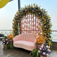 A beautiful floral arch for a Haldi or Mehendi ceremony, combining yellow flowers with hanging marigold strings and colorful tassels. The pink seashell-style sofa adds a touch of modern elegance to the vibrant, traditional setup.
