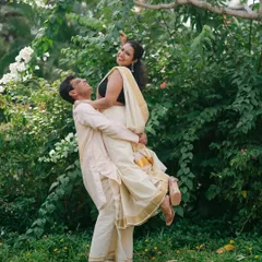 A playful and romantic moment as the groom lifts the bride, both dressed in elegant South Indian attire amidst a lush garden.