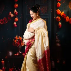 A graceful solo portrait celebrating tradition. The mom-to-be looks radiant in a classic cream and red saree, standing against a festive backdrop of flowers and candles.