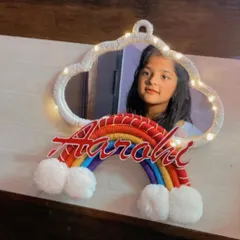 A happy young client with her new personalized 'Aarohi' cloud mirror. The mirror is framed with white macrame, features LED lights, and is decorated with a colorful rainbow name sign.
