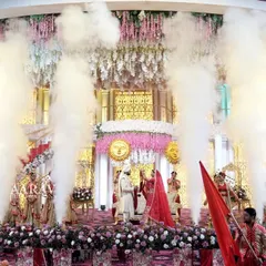 A wide shot of a grand Jaimala, showcasing the scale of the production. The couple is centered on a magnificent stage with smoke jets erupting around them, creating a breathtaking spectacle.