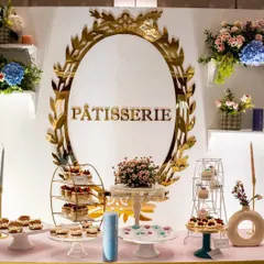 An exquisite "Pâtisserie" dessert table from a Parisian-themed baby shower. The display features a variety of delicate pastries on elegant stands, set against a white backdrop with a stunning gold-leaf mirror frame.