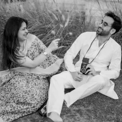 A playful and candid black and white moment. The couple shares a laugh in a field of tall grass, with the vintage camera prop adding a touch of old-school charm to their natural interaction.