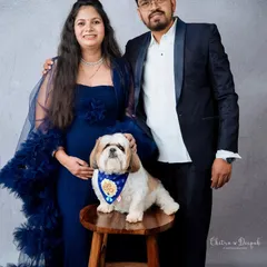 An elegant studio portrait of an expecting couple dressed in formal blue attire, with their well-behaved dog posing perfectly on a stool.