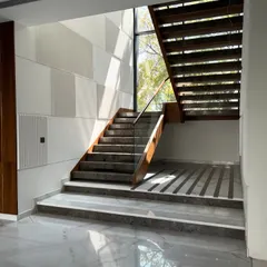 A view of the Gulmohar Farmhouse staircase, where the warmth of the wooden handrail and open risers contrasts with the cool, polished marble flooring. The striped pattern on the landing adds another layer of geometric detail.