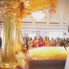 The couple stands before the Guru Granth Sahib during their Sikh wedding. This wide shot captures the entire scene, with family looking on in support.