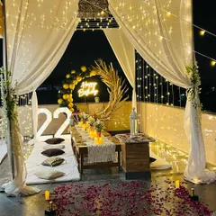 A complete view of my signature terrace cabana dinner setup for a 22nd birthday. This romantic arrangement includes a canopy with sheer white drapes and fairy lights, low seating with cushions, and rose petals scattered on the floor.