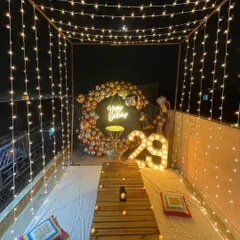 An intimate terrace birthday celebration for a 29th birthday. The setup includes a canopy of fairy lights, a balloon ring backdrop, marquee numbers, and a cozy low seating arrangement.