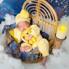 A peaceful newborn sleeps soundly in a yellow knit outfit, holding a tiny teddy bear. The setup includes a starry night backdrop, creating a dreamy, celestial feel.