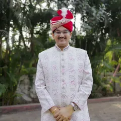 A confident and happy portrait of the groom in his elegant white sherwani and red turban (pheta), ready for his wedding day.