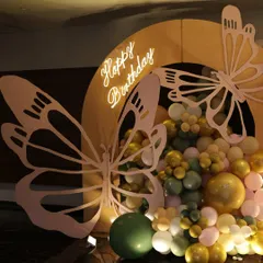 A close-up of our butterfly-themed birthday decor. This shot highlights the custom-cut wooden butterfly accent against a warm, arched backdrop and a neon "Happy Birthday" sign, nestled within a cascade of green and gold balloons.