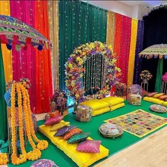 A vibrant and traditional Mehendi or Haldi seating area. The decor features a colorful backdrop of drapes, a floral ring, Rajasthani umbrellas, and low seating with bright cushions for a festive feel.