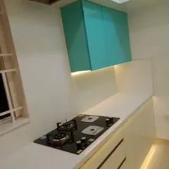This view focuses on the cooking zone, featuring an integrated four-burner hob set into the seamless white countertop. The teal overhead cabinets provide a pop of color, while the under-cabinet lighting ensures the workspace is well-lit for meal preparation.
