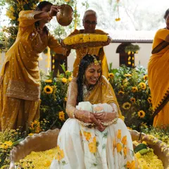 A beautiful moment during the Haldi ritual, where the bride is lovingly showered with flower petals and turmeric water by her family. The decor is filled with bright yellow flowers, enhancing the festive spirit.