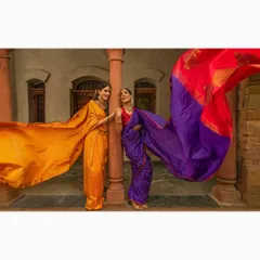 A fashion shoot featuring two models in vibrant silk sarees against an architectural backdrop. The wind catching the fabric creates a dramatic, flowing effect that showcases the beauty of the ethnic garments.