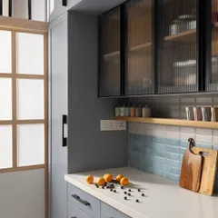 This compact kitchen blends pastel blue laminates with sleek black accents and warm wood finishes. A frosted glass sliding door separates the kitchen from the utility area, allowing natural light to flow through.