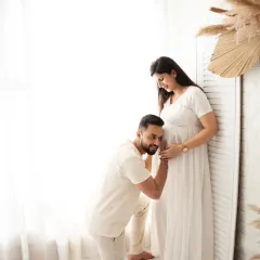 A classic pose filled with anticipation and love. The father-to-be kneels to listen to the baby bump, a tender moment captured in a beautifully lit, all-white studio setting.