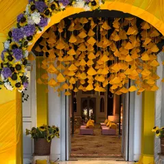 We love bringing vibrant traditions to life. This stunning Haldi ceremony entrance features a cascade of yellow fabric fans under an arch of bright sunflowers and purple blooms, welcoming guests into a joyous celebration.