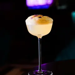 A sophisticated cocktail with a frothy top and delicate purple flower garnish, captured in a dark, moody lounge setting. The lighting highlights the elegant glassware and the drink's texture.