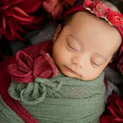 A close-up portrait focusing on the serene expression of this baby girl, surrounded by a bed of red flowers. The floral headband adds the perfect finishing touch.