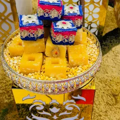 A modern and elegant sweet platter. Cubes of Mysore Pak are arranged on a bed of dal and topped with tiny decorative boxes, all presented on a silver plate and a golden stand.