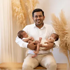 A proud father's smile says it all. He sits on a wooden bench in the studio, holding both of his newborn twins securely in his arms, beaming with happiness.