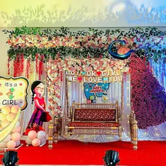 This gender reveal baby shower theme combines several popular elements. It features a central jhula, a stunning peacock made of flowers, a crescent moon prop, and 'It's a Boy/Girl' signs, all tied together with colorful lighting.