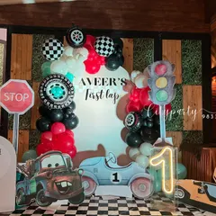 We brought the excitement of the racetrack to this 'First Lap' birthday celebration. The decor includes a custom arch backdrop, balloon garlands in red, black, and white, checkered flag details, tire props, and character cutouts from the movie 'Cars'.