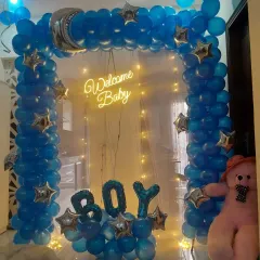 A "Welcome Baby Boy" decoration featuring a full arch of blue balloons, star accents, "BOY" foil letters, and a warm neon sign.