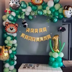 A jungle-themed first birthday decoration for a home setting. The balloon arch uses shades of green and is decorated with animal foil balloons and a birthday banner.