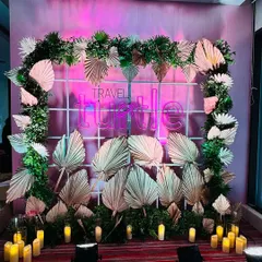 A chic boho-themed photo booth. The backdrop features a grid decorated with dried palm leaves and greenery, with a neon sign and candle lighting to create a trendy and inviting spot for photos.