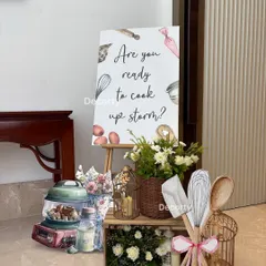 A charming welcome sign and decor vignette greets guests as they arrive. This setup uses rustic crates, fresh flowers, and oversized kitchen utensil props to immediately introduce the cooking theme.