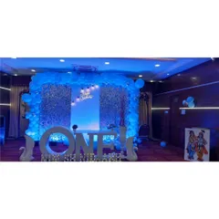 A stunning setup for a first birthday, featuring a large light-up 'ONE' sign as the centerpiece. The backdrop is a silver shimmer wall framed by a blue balloon arch, creating a cool, celebratory glow for the big milestone.