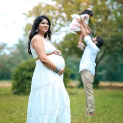 A beautiful outdoor maternity session capturing a candid family moment. The expectant mother is in focus, while in the background, the father joyfully lifts their older child in a sunlit park, telling a story of a growing family.
