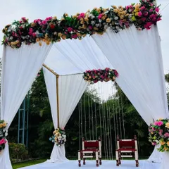 A simple and elegant outdoor wedding mandap with a white theme. The structure is draped with white fabric and adorned with colorful floral bouquets, perfect for a day wedding.