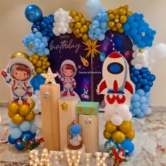 An outer space themed birthday party with a galaxy backdrop and astronaut cutouts. The setup includes wooden-style plinths, star props, and the child's name in marquee letters.