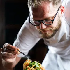 A chef proudly presenting a perfectly plated dish with tweezers. This shot captures the final moment of creation, highlighting the focus and artistry that goes into every plate in a fine-dining kitchen.