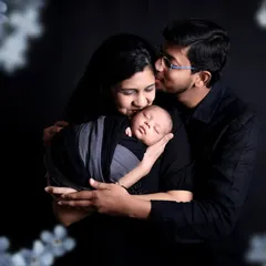 A tender family portrait where the father gently kisses his wife's forehead as they both hold their newborn baby. The dark background and soft floral overlay create a dreamy, romantic, and timeless feel.
