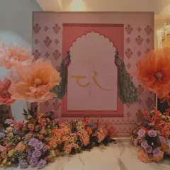 A beautiful home decoration with a custom TR monogram. The backdrop features a Rajasthani-style arch with peacocks, complemented by large peach and pink paper flowers.