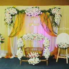 A beautiful setup for a prince's naming ceremony. The decor includes a flower-adorned cradle, a name reveal stand, and a backdrop of white and yellow drapes with lush floral arrangements.
