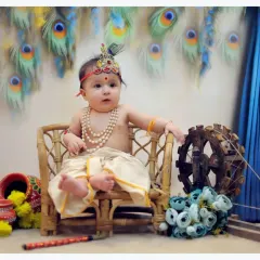 A variation of the Krishna theme, this shot features a baby sitting on a small stool with a spinning wheel prop. The background combines peacock feathers and marigold flowers for a vibrant look.