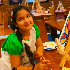 A young artist smiles proudly next to her butterfly painting. Our workshops provide all the materials and a supportive environment to let creativity flourish.
