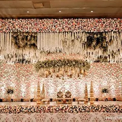 A breathtakingly large-scale stage design for a grand reception. The backdrop is a wall of countless flowers, with a floral chandelier and traditional golden seating, creating an opulent and luxurious atmosphere.